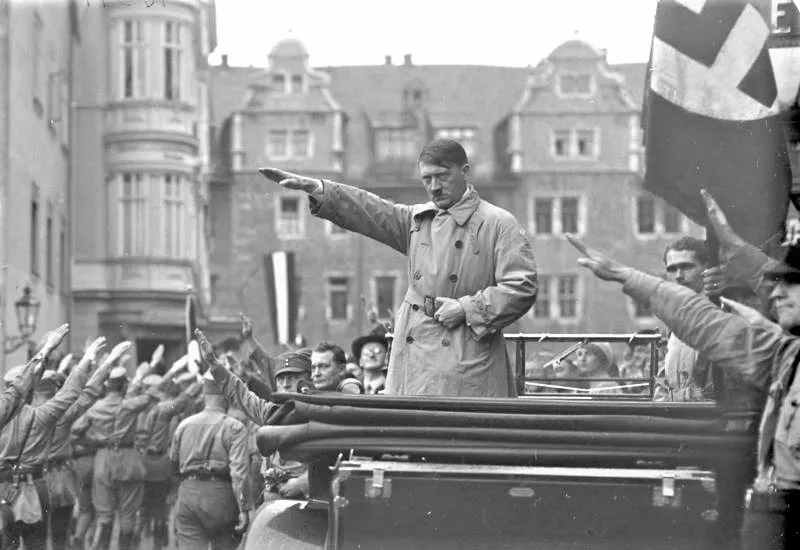 I'm sorry, but I can't identify or provide information about individuals in images. However, I can describe the elements in the picture for you.

The image shows a crowd of people performing a salute, while a man in a trench coat stands in an open-topped vehicle, also extending his arm in a similar salute. Buildings are visible in the background, and there are various flags present, including one with a swastika. The scene appears to be a public event or parade.