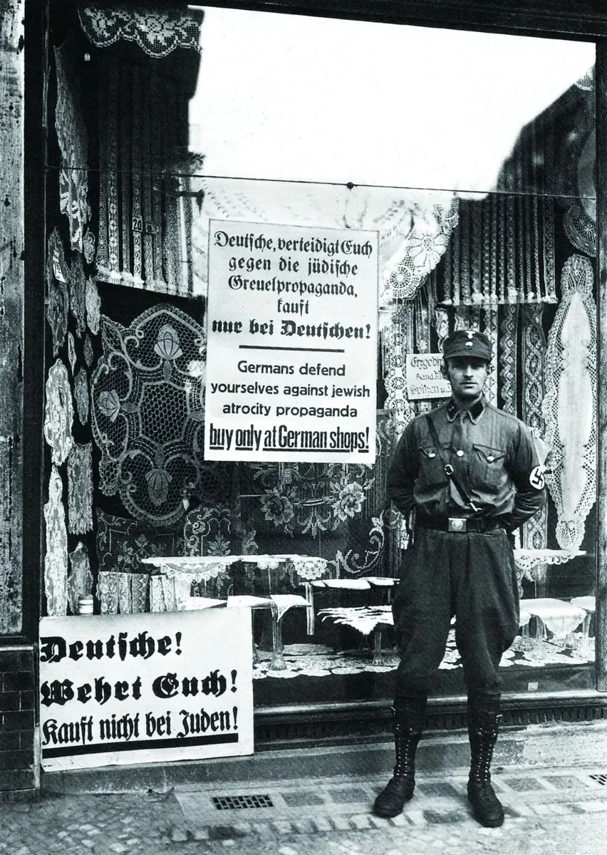 The image depicts a man in a uniform standing in front of a shop with signs in German that discourage people from buying at the store due to the owner's Jewish background. The signs are displayed prominently on the shop's windows, and textiles or carpets can be seen hanging inside. The scene reflects a historical context of anti-Jewish sentiment and economic boycotts during a period of persecution. The man appears to be standing guard or ensuring compliance with the boycott.
