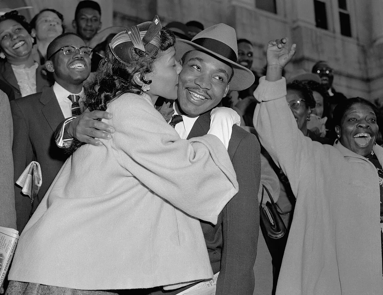 **Category 1: Clear and Distinct Elements and Symbolism**

The image captures a joyous moment involving Martin Luther King Jr. and his wife, Coretta Scott King. Martin Luther King Jr. is wearing a light-colored fedora hat and a dark suit with a tie. His expression is one of happiness and relief, evident by his broad smile showing his teeth. Coretta Scott King is embracing him warmly, planting a kiss on his cheek. She is dressed in a light-colored coat with a decorative hat featuring a bow, and her hair is styled in gentle waves.

In the background, a group of people surrounds them, all displaying expressions of happiness and excitement. Many faces are visible, with some individuals clapping or raising their hands in celebration. The crowd appears to be dressed in formal or semi-formal attire, typical of the mid-20th century, giving the impression of a significant event or gathering.

The image appears to have been taken outdoors in front of a large building, suggested by the visible stone façade and windows in the background. The photo is in black and white, highlighting the contrast between dark clothing and lighter elements like faces and coats.

**Category 2: Unclear and Interpretative Symbolism**

*The photograph captures what appears to be an event related to a moment of success or achievement, possibly linked to a court decision, given the historical context of Martin Luther King Jr.'s activism.*

*In the lower left corner, a newspaper is partly visible in another person's hand, hinting at the importance of current events during this time, though the text on the newspaper is unclear.*

*There is an air of solidarity and unity among the people, interpreting the raised hands and joyful expressions as possible signs of a communal victory or a shared success.*

The overall mood of the image is celebratory, reflecting a moment of triumph and intimacy between Martin Luther King Jr. and Coretta Scott King, while also capturing the collective joy of the community surrounding them.