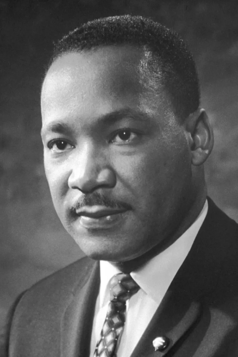 {
"title": "",
"description": "This black and white photograph prominently features Martin Luther King Jr., a well-known civil rights leader. \n\n**Category 1: Clearly Recognizable Elements and Symbolism**\n\n- **Pose and Expression:** Martin Luther King Jr. is shown from the shoulders upward, looking slightly to his right, which is to the viewer's left. His facial expression is composed and solemn, with a slight furrow in his brow, suggesting seriousness and contemplation. His eyes are wide open, focused, and convey a sense of determination and thoughtfulness.\n\n- **Clothing:** He is wearing a dark suit, with a white shirt and a patterned tie, which is visible in the image. His overall attire is formal, consistent with the dignified and respectful public persona he maintained.\n\n- **Background:** The background is blurred, which draws focus to his face. This lack of distraction emphasizes the importance of his expressions and features.\n\n**Category 2: Unclear, Interpretative Symbolism**\n\n- *Brooch or Pin:* A small pin or brooch is visible on his suit lapel. While it could symbolize membership or support for an organization, the details of this embellishment are indistinct, making it difficult to ascertain the precise symbolism.*\n\n- *Background Symbolism:* The blurred background could potentially represent historical or aspirational contexts, though this is an interpretative analysis.*\n\n**Additional Observations:**\n\n- **Lighting and Shading:** The lighting in the photograph creates subtle shadows on Martin Luther King Jr.'s face, particularly around his eyes and cheeks, adding depth and enhancing the serious tone of the image. The high contrast emphasizes his facial features and the texture of his hair and skin.\n\n- **Visual Focus:** The close-up nature of this photograph tightly focuses on his expression, accentuating the emotions and characteristics that made him a prominent and influential figure."
}