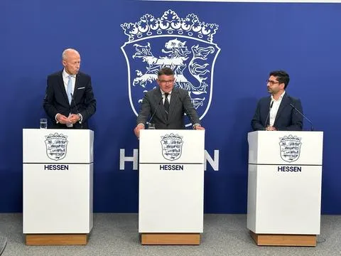 Pressevorstellung des hessischen Bildungspakets
Fotografie einer Pressevorstellung in der Hessischen Staatskanzlei: Kultusminister Armin Schwarz, Ministerpräsident Boris Rhein und Vize-Ministerpräsident Kaweh Mansoori stellen das Bildungspaket vor.

--- Image Analysis ---
1. **Data & Statistics**: 
   - 3 politicians/individuals are visible standing behind separate podiums.

2. **Text Content**:
   - "HESSEN" (written in capital letters on each of the three white podiums, accompanied by the state's coat of arms above the text).

3. **Visual Structure**:
   - A photograph showing a press conference or official government statement setup.
   - Background: A blue wall featuring a large white outline of the Hessian coat of arms (Hessischer Löwe) and partially visible white lettering (part of "HESSEN").
   - Foreground: Three identical white podiums with wooden bases. Each podium displays the state crest of Hessen and the word "HESSEN".
   - Subjects: Three men in suits standing behind the podiums, each with a microphone.

4. **Key Takeaways**:
   - The image depicts an official press conference or political statement involving three representatives of the state of Hessen.
   - Official state branding, including the Hessian coat of arms and the name "HESSEN", is prominently featured on both the backdrop and the podiums.