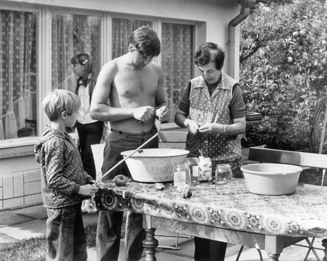 Alltag in der DDR: Obst einwecken
Das Bild zeigt eine typische Alltagsszene in der DDR. Aufgrund von Versorgungsengpässen und um für den Winter vorzusorgen, war das Einwecken von Obst und Gemüse aus dem eigenen Garten (falls vorhanden) weit verbreitet. Es symbolisiert die Notwendigkeit zur Selbstversorgung in der Mangelwirtschaft.
--- Image Analysis ---
Here's the analysis of the image, broken down into the requested categories:
1. **Data & Statistics:**
* None directly visible in the image.
2. **Text Content:**
* There is no text visible in the image.
3. **Visual Structure:**
* The image is a black and white photograph showing a family preparing food, likely preserving or canning fruits or vegetables. A man (possibly the father) is shirtless and seems to be peeling something. A woman (possibly the mother) is also preparing food and wearing an apron. A young boy is standing at the table. There is a girl standing behind the boy. A table with a patterned tablecloth is set up outdoors, holding bowls, jars, and what appears to be the produce they are working with. There is a house with windows in the background, as well as a tree.
4. **Key Takeaways:**
* The image depicts a family engaged in a food preservation activity, possibly during a harvest season.
* The scene likely represents a period or cultural practice of preserving food at home.