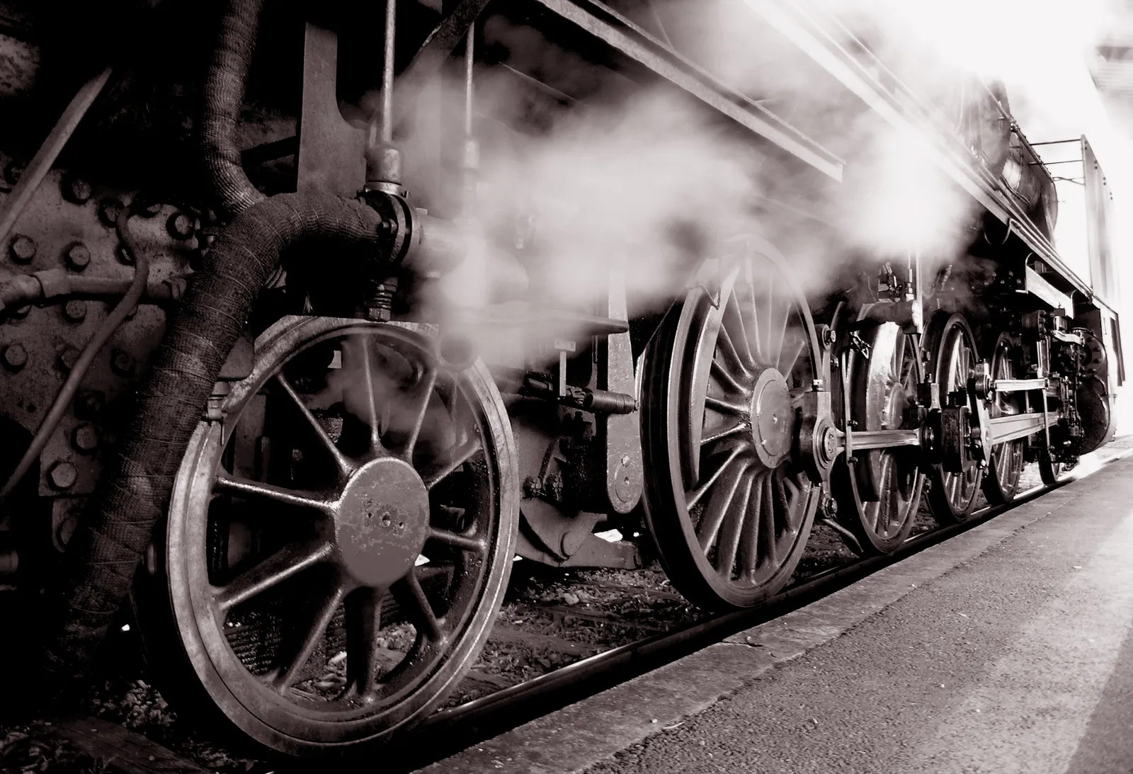 Dampflokomotive in Bewegung
Nahaufnahme einer dampfbetriebenen Lokomotive mit großen Treibrädern, Gestänge und austretendem Dampf. Das Bild dient als visuelle Orientierung für die Technik-Station.
--- Image Analysis ---
1. **Data & Statistics**: None (the image contains no numbers, percentages, dates, or statistical data points).
2. **Text Content**: None (the image contains no visible text, labels, headings, or captions).
3. **Visual Structure**:
- This is a close-up, black-and-white photograph depicting the side and undercarriage of a historic steam locomotive.
- The perspective is angled from low to high, focusing heavily on the large, spoked metal driving wheels resting on the rails next to a station platform.
- Thick plumes of steam and vapor are captured emerging around the machinery and wheels, creating a dynamic, atmospheric effect.
- The complex arrangement of iron rods, pistons, pipes, and riveted metal plates of the locomotive body is clearly visible.
4. **Key Takeaways**:
- The image visually demonstrates the heavy iron machinery and complex mechanical systems (such as pistons, rods, and large spoked wheels) characteristic of historical steam locomotives.
- It captures the iconic production of steam and vapor that powered rail transport during the industrial era.