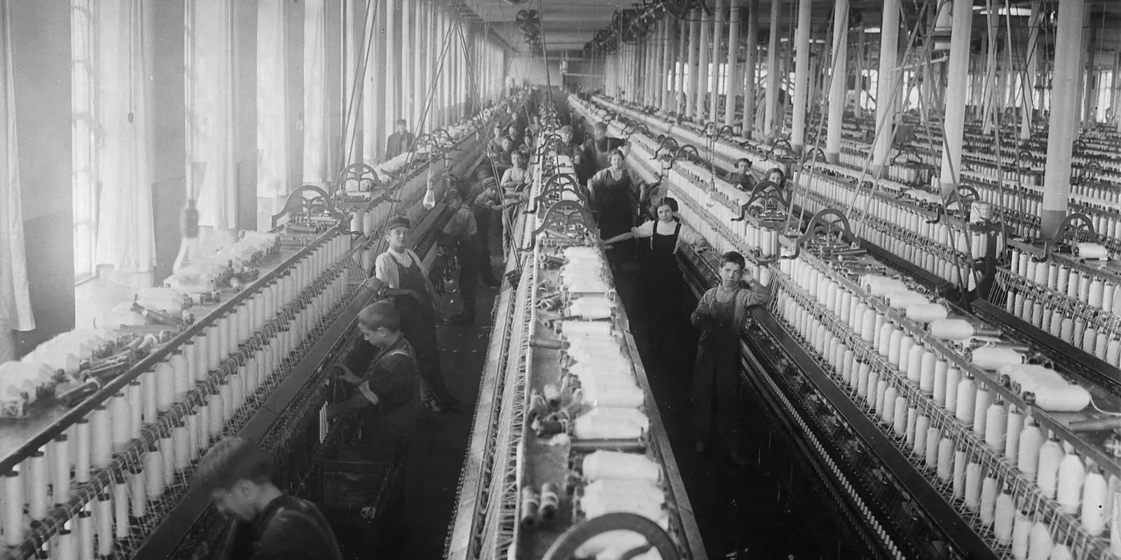 Arbeit in einer Textilfabrik
Historische Schwarzweißaufnahme einer großen Textilfabrik mit langen Reihen von Maschinen und arbeitenden Personen. Die Aufnahme stammt aus den USA und dient als Vergleichsbild für industrialisierte Fabrikarbeit.
--- Image Analysis ---
1. **Data & Statistics**: Keine expliziten Daten oder Statistiken sichtbar.
2. **Text Content**: Keine sichtbaren Texte, Überschriften oder Beschriftungen im Bild.
3. **Visual Structure**: Es handelt sich um ein historisches Schwarz-Weiß-Foto, das das Innere einer Textilfabrik (Spinnerei) zeigt. Lange Reihen von industriellen Spinnmaschinen erstrecken sich durch den tiefen Raum, begleitet von vielen Arbeiterinnen und Arbeitern, darunter auch erkennbar jugendliche Arbeitskräfte (Kinderarbeit während der Industrialisierung). Die Maschinen sind voll mit Spulen und Garnen, während im Hintergrund große Fenster viel Tageslicht hereinlassen.
4. **Key Takeaways**:
- Das Bild illustriert die Arbeitsbedingungen während der Industrialisierung, insbesondere die Massenproduktion von Textilien in großen Fabriken.
- Es zeigt den historischen Einsatz von Kinderarbeit in Fabriken, wo Minderjährige oft gefährliche oder monotone Tätigkeiten an Maschinen verrichten mussten.
- Die Fotografie verdeutlicht die Mechanisierung und die langen Reihen von Maschinen (Spinnmaschinen), die für die Fabrikarbeit im 19. oder frühen 20. Jahrhundert typisch waren.