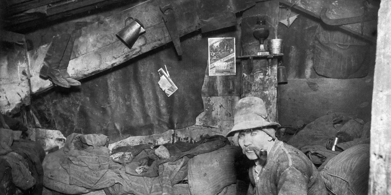 Beengte Wohnung in einem Mietshaus
Historische Schwarzweißaufnahme eines sehr kleinen, vollgestellten Innenraums mit mehreren Personen. Die Aufnahme entstand im späten 19. Jahrhundert in New York und dient als Vergleichsbild für Wohnungsnot und Enge in Industriestädten.
--- Image Analysis ---
1. **Data & Statistics**:
- The visible calendar page shows the year **1891** and the month **January**.
2. **Text Content**:
- On the calendar hanging on the wall: "JANUARY 1891" and the name "S. ZUSCHLAG".
3. **Visual Structure**:
- A historical black-and-white interior photograph showing a cramped, rudimentary dwelling or shack.
- A man wearing a hat and smoking a pipe is seated in the foreground, looking directly at the camera.
- The walls and ceiling are made of rough wood and tar paper or fabric. Various items are hung or stored on the walls and beams: boots, a metal container/jug, a hand saw, a small calendar, a small shelf with a kerosene lamp and cup, and fabric bags.
- The sleeping/living area consists of piles of blankets and straw sacks filling the background and sides of the small room.
4. **Key Takeaways**:
- The image depicts the extremely poor living conditions and impoverished housing common among certain working-class people or laborers in the late 19th century.
- The cramped, makeshift interior reflects cramped urban living conditions or temporary shelters (such as lodging houses or tenements) during the industrial era.