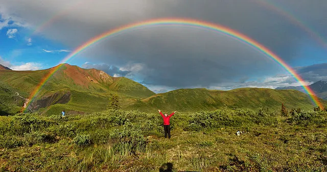 Material D: Regenbogen nach dem Sturm
Ein farbenfroher doppelter Regenbogen spannt sich über eine weite, grüne Landschaft nach einem Unwetter. Der Himmel ist noch teilweise von dunklen Wolken bedeckt, aber die Sonne bricht bereits durch.

--- Image Analysis ---
This image is a simple photograph with no data, statistics, or text content.

1.  **Data & Statistics**: None visible.
2.  **Text Content**: None visible.
3.  **Visual Structure**: The image is a wide panoramic photograph of a natural landscape. It features a prominent, vibrant rainbow arching across the sky, with a fainter secondary rainbow visible above it. The foreground consists of green, bushy vegetation and grassy terrain. In the mid-ground, there are rolling green hills and mountains, with some reddish-brown patches on one of the peaks. A small waterfall or stream can be seen flowing down one of the hills on the left. The sky is a mix of bright blue with white clouds on the left and dark, stormy grey clouds on the right, indicating recent or ongoing rain. Two human figures are visible in the landscape: one in the center wearing a red jacket with arms outstretched, and another smaller figure in blue further to the left. A small white dog is also visible near the person in red.
4.  **Key Takeaways**:
    *   Rainbows are natural optical phenomena that occur when sunlight shines through water droplets, typically after rain.
    *   Sometimes, a secondary, fainter rainbow can be observed outside the primary rainbow, with the colors reversed.
    *   Natural landscapes can offer breathtaking views, especially when combined with atmospheric phenomena like rainbows.