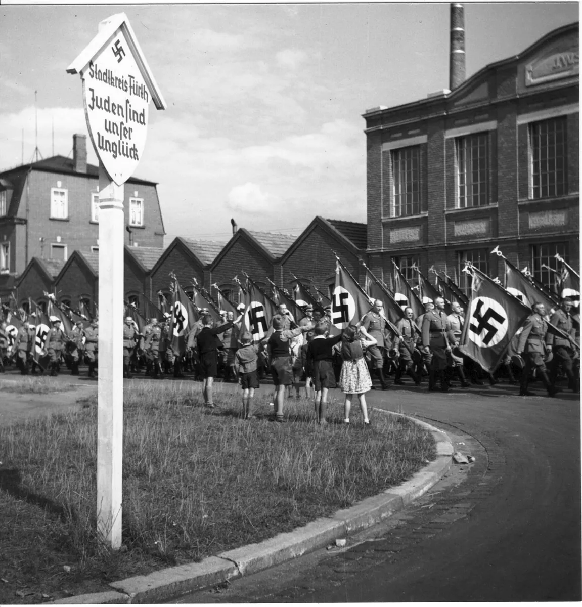 NS-Marschkolonne mit Hakenkreuzfahnen auf dem Rückweg vom Reichsparteitag (vermutlich 1938) an der Stadtgrenze Fürth/Nürnberg, antijüdische Propaganda am Ortsschild und Kinder mit dem (zu jener Zeit vor NS-Fahnen vorgeschriebenen) Hitlergruß.[1] Im Hintergrund ein Fabrikgebäude der „arisierten“, zuvor jüdischen Firma J.W. Spear & Söhne. Im Vordergrund ein Schild mit der Aufschrift „Stadtkreis Fürth / Juden sind unser Unglück“