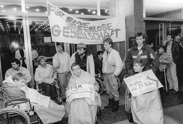 Berlin, Januar 1990: Rollstuhlfahrer demonstrieren vor einem Kino. Unter der Losung „Gegen bauliche und geistige Barrieren – für zugängliche Menschen und Gebäude“ forderten sie mit ihrer 20-minütigen Blockade des Kinoeingangs, Behinderte nicht länger vom kulturellen Leben auszugrenzen.